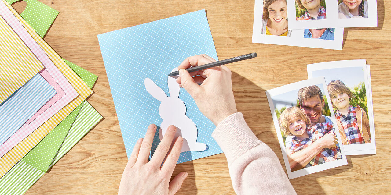 Dvě ruce kreslí velikonočního zajíčka na papír pomocí šablony. Vedle něj jsou vytištěné okamžité fotografie rodiny.